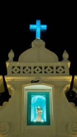 A religious structure with a lit blue cross at the top against a dark sky. Below the cross is a small alcove containing a statue of a saint, illuminated in a turquoise light. The facade of the building is ornate with decorative elements and has a classic architectural style.