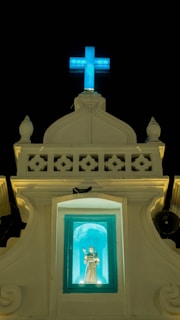 A religious structure with a lit blue cross at the top against a dark sky. Below the cross is a small alcove containing a statue of a saint, illuminated in a turquoise light. The facade of the building is ornate with decorative elements and has a classic architectural style.
