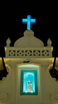 A religious structure with a lit blue cross at the top against a dark sky. Below the cross is a small alcove containing a statue of a saint, illuminated in a turquoise light. The facade of the building is ornate with decorative elements and has a classic architectural style.