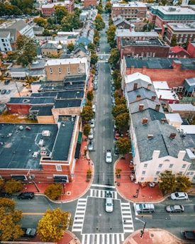 An aerial view of a small urban area featuring a straight road lined with parked cars and bordered by buildings. The architecture consists of both modern and older brick structures. There are several trees along the sidewalks, providing some greenery amid the urban landscape. The road and pedestrian crosswalks are clearly visible, indicating a quiet and organized neighborhood.