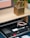 A wooden desk holds a golden sculptural piece. Below the desk, a drawer contains several electronic accessories organized neatly. These include a USB drive, cables, a Samsung SSD, a memory card, and other small adapters.