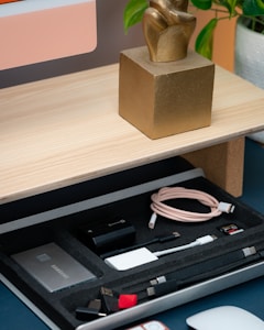 A wooden desk holds a golden sculptural piece. Below the desk, a drawer contains several electronic accessories organized neatly. These include a USB drive, cables, a Samsung SSD, a memory card, and other small adapters.