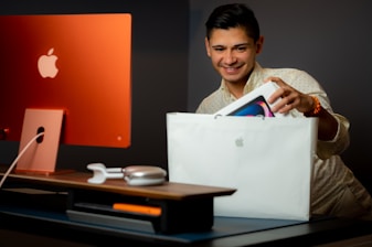 A person with short dark hair is smiling while holding a white Apple shopping bag and taking out a boxed Apple product. The setting includes a stylish workspace with an orange iMac computer and accessories like headphones and a mouse on a desk.