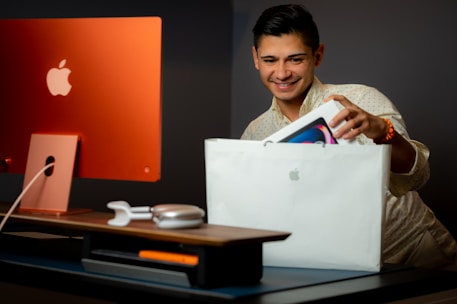 A person with short dark hair is smiling while holding a white Apple shopping bag and taking out a boxed Apple product. The setting includes a stylish workspace with an orange iMac computer and accessories like headphones and a mouse on a desk.
