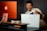 A person with short dark hair is smiling while holding a white Apple shopping bag and taking out a boxed Apple product. The setting includes a stylish workspace with an orange iMac computer and accessories like headphones and a mouse on a desk.
