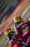 Two motocross riders wearing vibrant helmets and matching red and black racing suits are captured mid-action. They are leaning into what appears to be a sharp turn on a dirt track, with the motion creating a blur of the background.