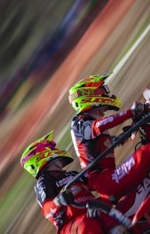 Two motocross riders wearing vibrant helmets and matching red and black racing suits are captured mid-action. They are leaning into what appears to be a sharp turn on a dirt track, with the motion creating a blur of the background.