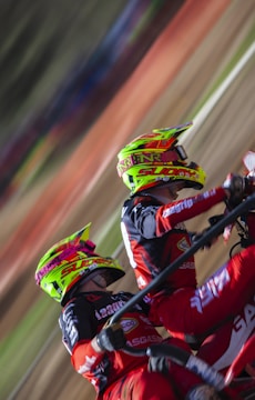 Two motocross riders wearing vibrant helmets and matching red and black racing suits are captured mid-action. They are leaning into what appears to be a sharp turn on a dirt track, with the motion creating a blur of the background.