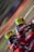 Two motocross riders wearing vibrant helmets and matching red and black racing suits are captured mid-action. They are leaning into what appears to be a sharp turn on a dirt track, with the motion creating a blur of the background.
