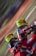Two motocross riders wearing vibrant helmets and matching red and black racing suits are captured mid-action. They are leaning into what appears to be a sharp turn on a dirt track, with the motion creating a blur of the background.