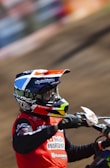 A person wearing a colorful helmet and red racing jersey is riding a motocross bike. The background is a blurred dirt track, suggesting motion and speed. The person's gear includes sponsor logos and intricate designs on the helmet.
