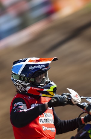 A person wearing a colorful helmet and red racing jersey is riding a motocross bike. The background is a blurred dirt track, suggesting motion and speed. The person's gear includes sponsor logos and intricate designs on the helmet.