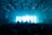 A crowded concert venue with a large audience in front of a stage. A DJ is silhouetted against bright blue and white light displays. The ceiling is filled with metal frameworks and spotlights pointing toward the audience. People are raising their hands and cameras, capturing the energetic atmosphere.