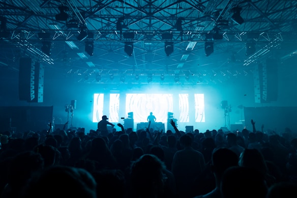 A crowded concert venue with a large audience in front of a stage. A DJ is silhouetted against bright blue and white light displays. The ceiling is filled with metal frameworks and spotlights pointing toward the audience. People are raising their hands and cameras, capturing the energetic atmosphere.