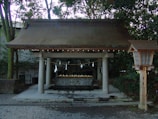 A traditional Japanese purification pavilion with wooden roof and stone basin, typically found at Shinto shrines. The structure is surrounded by dense greenery, and white paper decorations hang from a rope above the basin, which is used for ritual cleansing. A wooden lantern stands to the right, adding to the authentic cultural setting.
