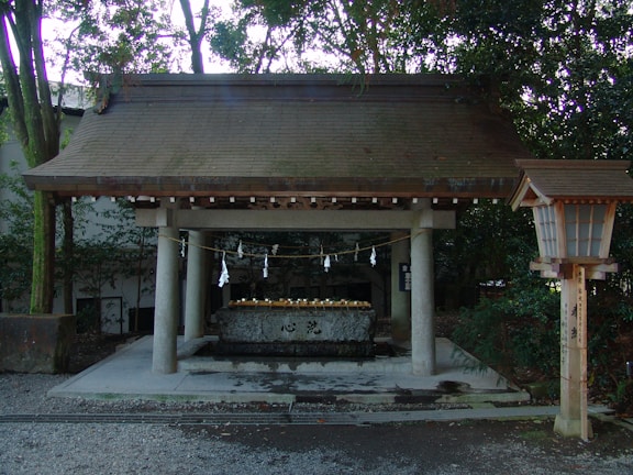 A traditional Japanese purification pavilion with wooden roof and stone basin, typically found at Shinto shrines. The structure is surrounded by dense greenery, and white paper decorations hang from a rope above the basin, which is used for ritual cleansing. A wooden lantern stands to the right, adding to the authentic cultural setting.