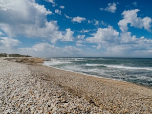 a rocky beach next to the ocean under a cloudy blue sky