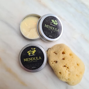 Metal tins labeled 'Mendula Face Cream' and 'Mendula Hand Cream' sit open on a light marble surface. A natural sponge is positioned nearby, contributing a soft, organic texture to the arrangement. The face cream tin is slightly open, revealing a smooth, creamy substance inside.