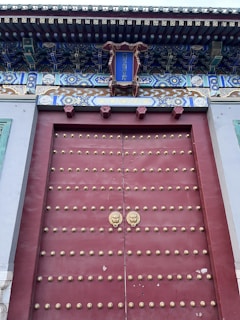 A large traditional wooden door, painted red and adorned with rows of gold-colored studs. There are two ornate door knockers shaped like lion heads. Above the door, there is a decorative panel with intricate blue, green, and gold patterns, with a sign in the center featuring Chinese characters. The overhang above the door includes elaborate woodwork with detailed carvings and painting.