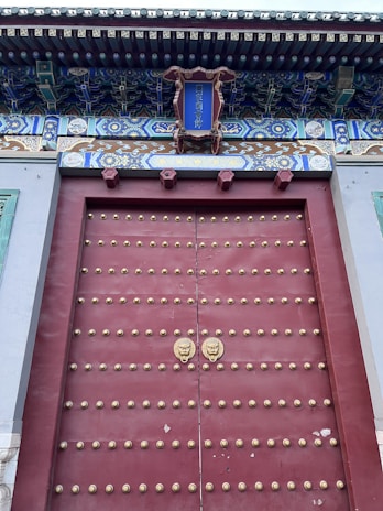 A large traditional wooden door, painted red and adorned with rows of gold-colored studs. There are two ornate door knockers shaped like lion heads. Above the door, there is a decorative panel with intricate blue, green, and gold patterns, with a sign in the center featuring Chinese characters. The overhang above the door includes elaborate woodwork with detailed carvings and painting.