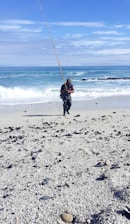 A person is standing on a sandy beach holding a long fishing rod. The ocean waves are gently crashing behind, and the sky is clear with some clouds. The foreground is covered in pebbles and small shells scattered across the sand.