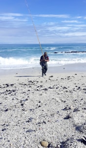 A person is standing on a sandy beach holding a long fishing rod. The ocean waves are gently crashing behind, and the sky is clear with some clouds. The foreground is covered in pebbles and small shells scattered across the sand.