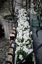 A row of white flowers is beautifully arranged in a rectangular planter, placed on a stone surface. The planter has metal embellishments resembling hooks. Surrounding the planter are rustic elements, including a woven basket and branches contained in a large glass vase. The background includes cobblestone and a white column.