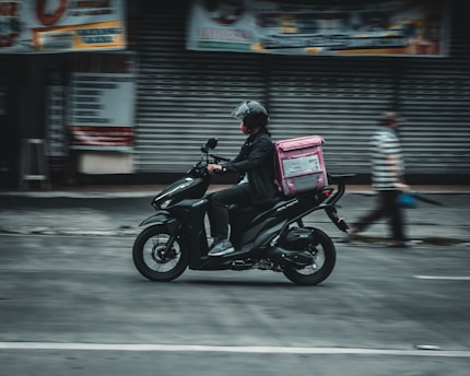 A person wearing a helmet rides a black scooter on a street, carrying a pink delivery bag on the back. The background features a closed shop with metallic shutters, and a blurred pedestrian walks nearby, suggesting motion and urban setting.