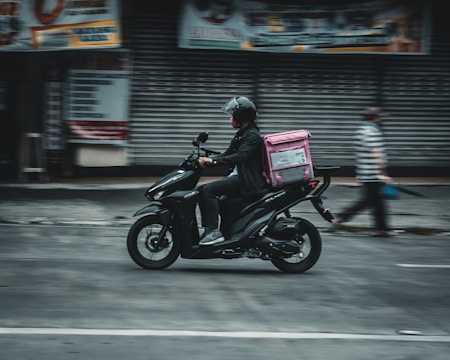 A person wearing a helmet rides a black scooter on a street, carrying a pink delivery bag on the back. The background features a closed shop with metallic shutters, and a blurred pedestrian walks nearby, suggesting motion and urban setting.
