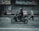 A person wearing a helmet rides a black scooter on a street, carrying a pink delivery bag on the back. The background features a closed shop with metallic shutters, and a blurred pedestrian walks nearby, suggesting motion and urban setting.