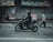 A person wearing a helmet rides a black scooter on a street, carrying a pink delivery bag on the back. The background features a closed shop with metallic shutters, and a blurred pedestrian walks nearby, suggesting motion and urban setting.