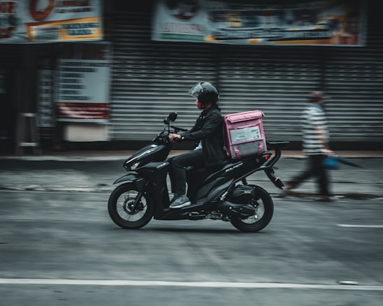 A person wearing a helmet rides a black scooter on a street, carrying a pink delivery bag on the back. The background features a closed shop with metallic shutters, and a blurred pedestrian walks nearby, suggesting motion and urban setting.