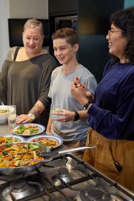 Three people stand around a kitchen stove filled with colorful cooking dishes. Each person holds a drink, laughing and enjoying each other's company. The setting suggests a friendly and informal cooking session.
