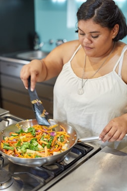 A woman is cooking in a kitchen, stirring vibrant, chopped vegetables in a frying pan on a gas stove. She is wearing a white tank top and a pendant necklace.