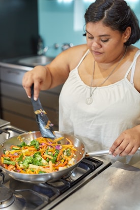 A woman is cooking in a kitchen, stirring vibrant, chopped vegetables in a frying pan on a gas stove. She is wearing a white tank top and a pendant necklace.