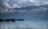 A serene lake is surrounded by lush greenery and distant mountains shrouded in mist and clouds. Several houseboats float gently on the calm water, which reflects the sky and surrounding landscape. The atmosphere is peaceful and tranquil, with a sense of stillness.