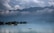A serene lake is surrounded by lush greenery and distant mountains shrouded in mist and clouds. Several houseboats float gently on the calm water, which reflects the sky and surrounding landscape. The atmosphere is peaceful and tranquil, with a sense of stillness.
