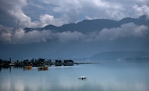 A serene lake is surrounded by lush greenery and distant mountains shrouded in mist and clouds. Several houseboats float gently on the calm water, which reflects the sky and surrounding landscape. The atmosphere is peaceful and tranquil, with a sense of stillness.