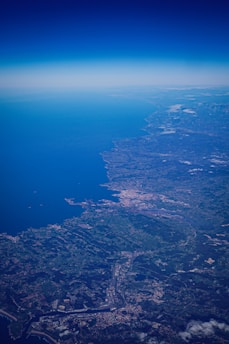 An aerial view of a coastal region with a large body of water extending to the horizon, adjacent to sprawling urban and rural landscapes. Small clouds are scattered across the landscape and city formations are visible near the shoreline.