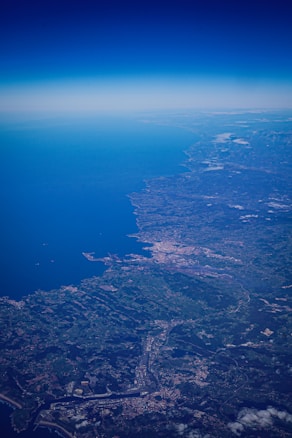 An aerial view of a coastal region with a large body of water extending to the horizon, adjacent to sprawling urban and rural landscapes. Small clouds are scattered across the landscape and city formations are visible near the shoreline.