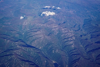 An aerial view of rugged mountainous terrain with ridges and valleys clearly visible. Sparse clouds float above the landscape, casting shadows on the undulating surface below. The area features a mix of rocky outcrops and patches of vegetation, with variations in elevation highlighted by shadows.