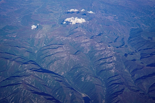 An aerial view of rugged mountainous terrain with ridges and valleys clearly visible. Sparse clouds float above the landscape, casting shadows on the undulating surface below. The area features a mix of rocky outcrops and patches of vegetation, with variations in elevation highlighted by shadows.