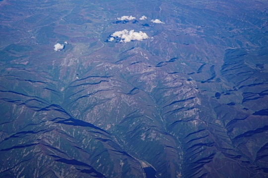 An aerial view of rugged mountainous terrain with ridges and valleys clearly visible. Sparse clouds float above the landscape, casting shadows on the undulating surface below. The area features a mix of rocky outcrops and patches of vegetation, with variations in elevation highlighted by shadows.