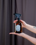 A pair of hands is holding a brown glass spray bottle with a black nozzle. The bottle has a minimalist label with a floral design and the words 'press & breathe'. The background features a dark curtain, and the person has painted nails and a tattoo on their forearm.