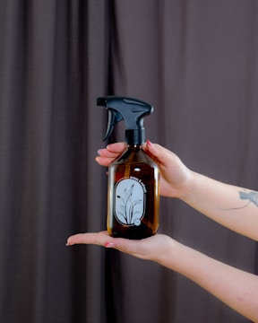 A pair of hands is holding a brown glass spray bottle with a black nozzle. The bottle has a minimalist label with a floral design and the words 'press & breathe'. The background features a dark curtain, and the person has painted nails and a tattoo on their forearm.