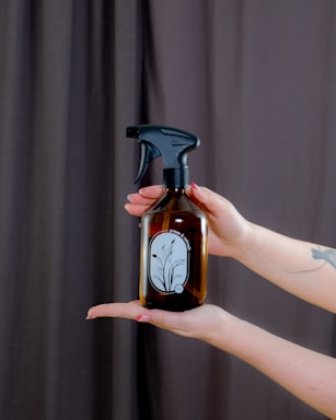 A pair of hands is holding a brown glass spray bottle with a black nozzle. The bottle has a minimalist label with a floral design and the words 'press & breathe'. The background features a dark curtain, and the person has painted nails and a tattoo on their forearm.