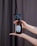 A pair of hands is holding a brown glass spray bottle with a black nozzle. The bottle has a minimalist label with a floral design and the words 'press & breathe'. The background features a dark curtain, and the person has painted nails and a tattoo on their forearm.