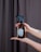 A pair of hands is holding a brown glass spray bottle with a black nozzle. The bottle has a minimalist label with a floral design and the words 'press & breathe'. The background features a dark curtain, and the person has painted nails and a tattoo on their forearm.