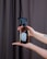 A pair of hands is holding a brown glass spray bottle with a black nozzle. The bottle has a minimalist label with a floral design and the words 'press & breathe'. The background features a dark curtain, and the person has painted nails and a tattoo on their forearm.