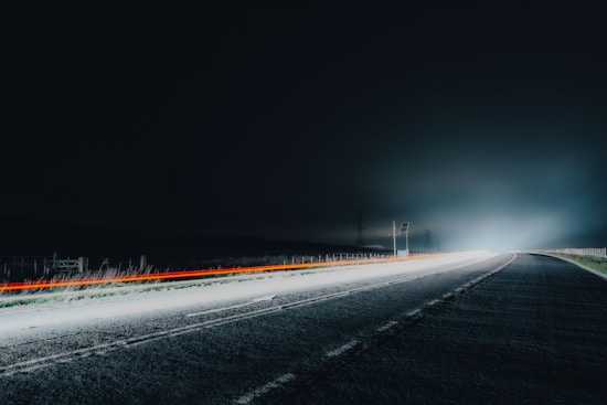 A long stretch of empty road at night illuminated by bright headlights, creating streaks of light. The road is surrounded by darkness, with faint silhouettes of fencing and poles on either side.